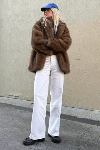 model-wearing-camille-coat-golden-brown-faux-fur-arms-crossed-styled-with-white-wide-leg-jeans-and-blue-cap