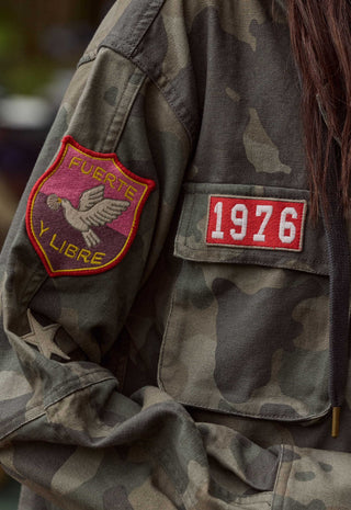 Close-up of a camouflage military-style jacket with a red and yellow bird patch on the sleeve and a red rectangular patch with '1976' on the chest pocket.