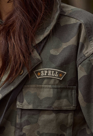 Close-up of a person wearing a camouflage utility jacket, showing the chest pocket with a sewn 'SPELL' patch on the left chest.